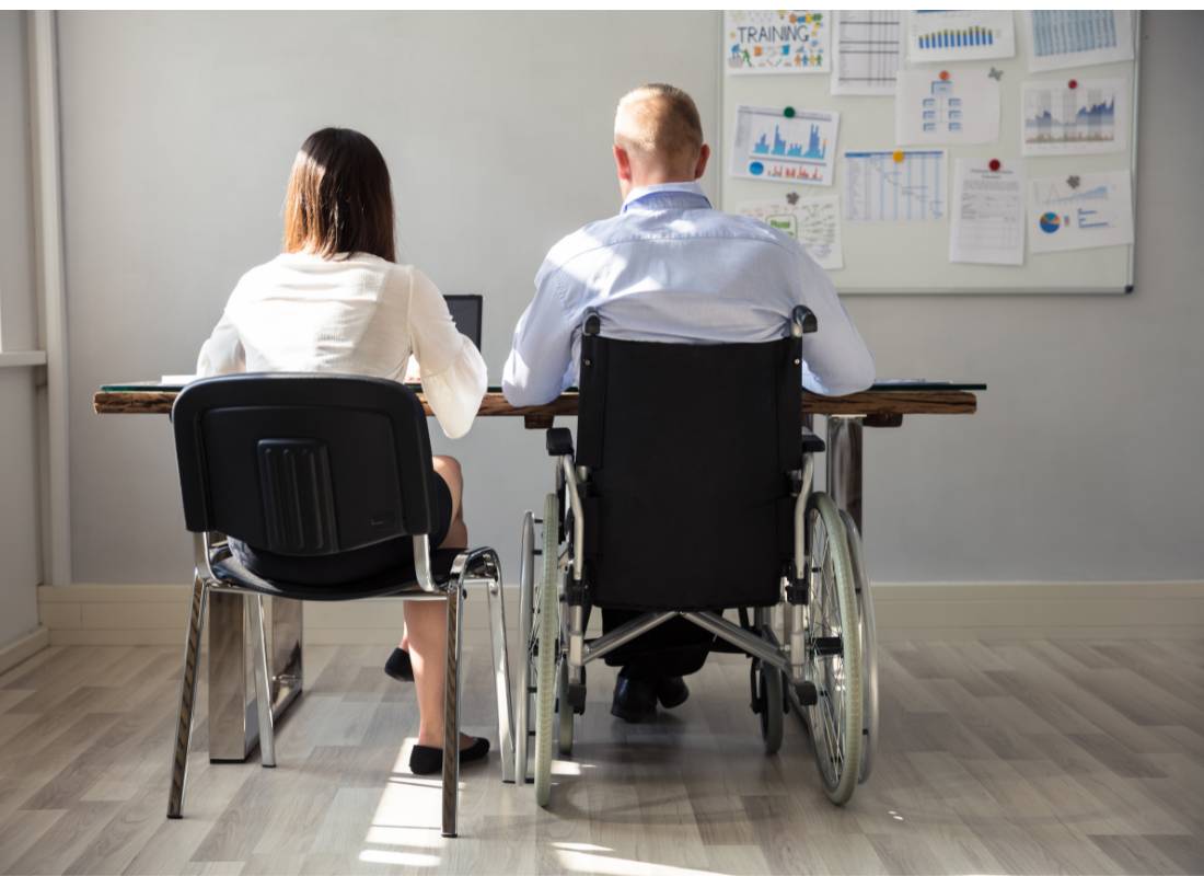 Mężczyzna na wózku pracuje z kobietą przy jednym biurku. Obrazek w artykule Praca z osobami niepełnosprawnymi